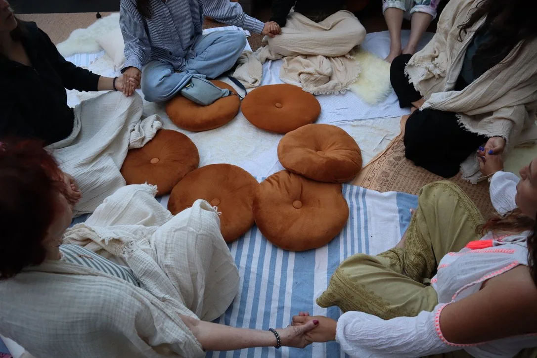 A circle of people seated on cushions on the floor, holding hands