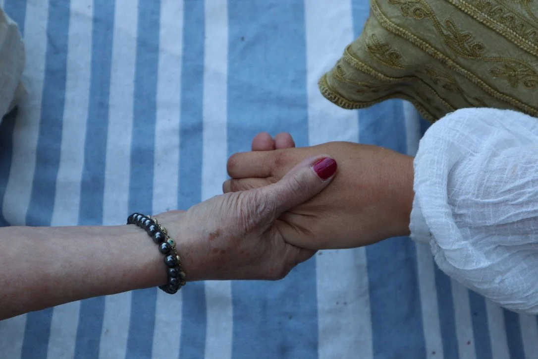 Two hands holding each other, one with a black and silver beaded bracelet and the other with painted nails, over a blue and white striped fabric background.